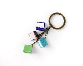Op de afbeelding: Een sleutelhanger met vier miniatuurboeken in groen, turquoise, bordeauxrood en paars. De kleine boeken zijn bevestigd aan een bronzen sleutelring met een ketting. De boeken hebben witte pagina's.