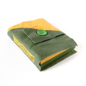 May include: A handmade leather wallet in shades of olive green and mustard yellow. The wallet is secured with a green button closure. The design includes a stitched yellow pattern on the front. The wallet is likely for carrying cards or cash.