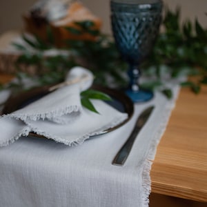 May include: A white linen table runner with frayed edges, a black plate with a white napkin, a silver knife, and a blue glass on a wooden table.