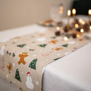 Può includere: Runner da tavolo natalizio beige con motivi festivi. Presenta omini di pan di zenzero, pupazzi di neve, alberi di Natale e fiocchi di neve. Il runner è posizionato su una tovaglia bianca.