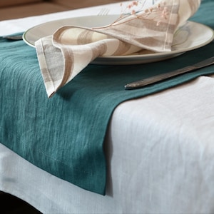 May include: A teal linen table runner with a white tablecloth underneath. A white plate with a striped napkin and a sprig of dried flowers sits on top. A silver knife is placed to the side, creating a simple, elegant table setting.