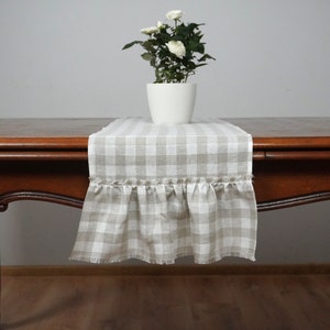 Puede incluir: Un camino de mesa a cuadros beige y blanco con un borde de volantes. El camino de mesa está hecho de lino y tiene un borde deshilachado. El camino de mesa está sobre una mesa de madera con un arreglo de flores blancas en una maceta blanca.