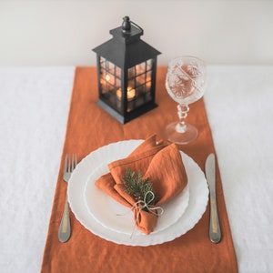 May include: A white dinner plate with a folded orange linen napkin tied with twine and a sprig of greenery. The plate is set on a brown linen table runner with a black lantern and a clear glass wine glass.
