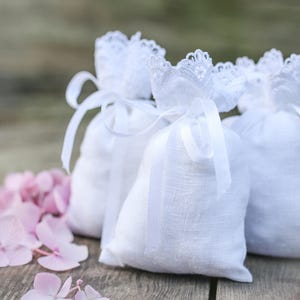 May include: Three white linen sachets with lace trim and ribbon ties, arranged on a wooden surface with pink hydrangea flowers. The sachets are filled and tied with white ribbons.