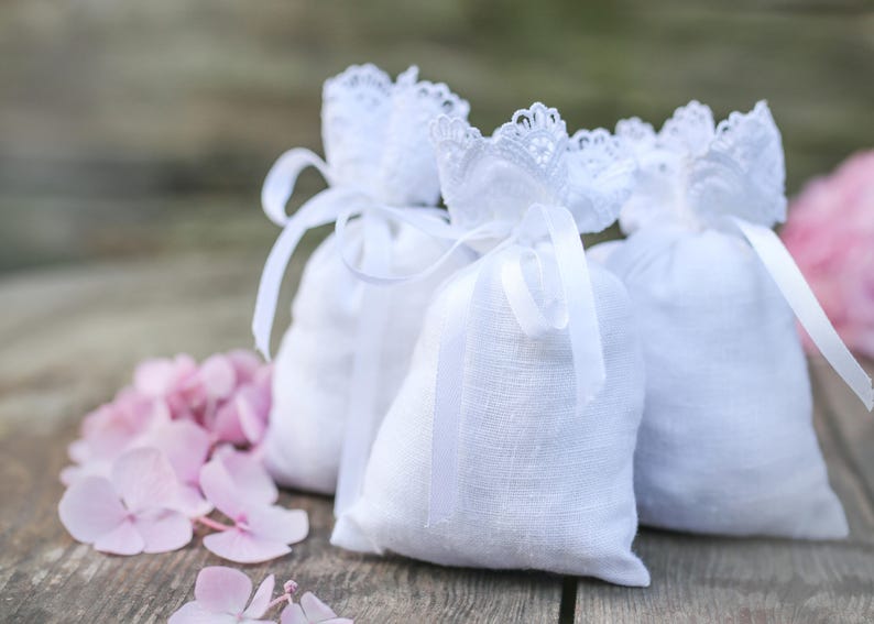 May include: Three white linen sachets with lace trim and ribbon ties, arranged on a wooden surface with pink hydrangea flowers. The sachets are filled and tied with white ribbons.