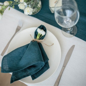 Puede incluir: Una servilleta de lino azul verdoso doblada en forma de triángulo y colocada sobre un plato blanco. Una sola flor blanca está metida en el pliegue de la servilleta.