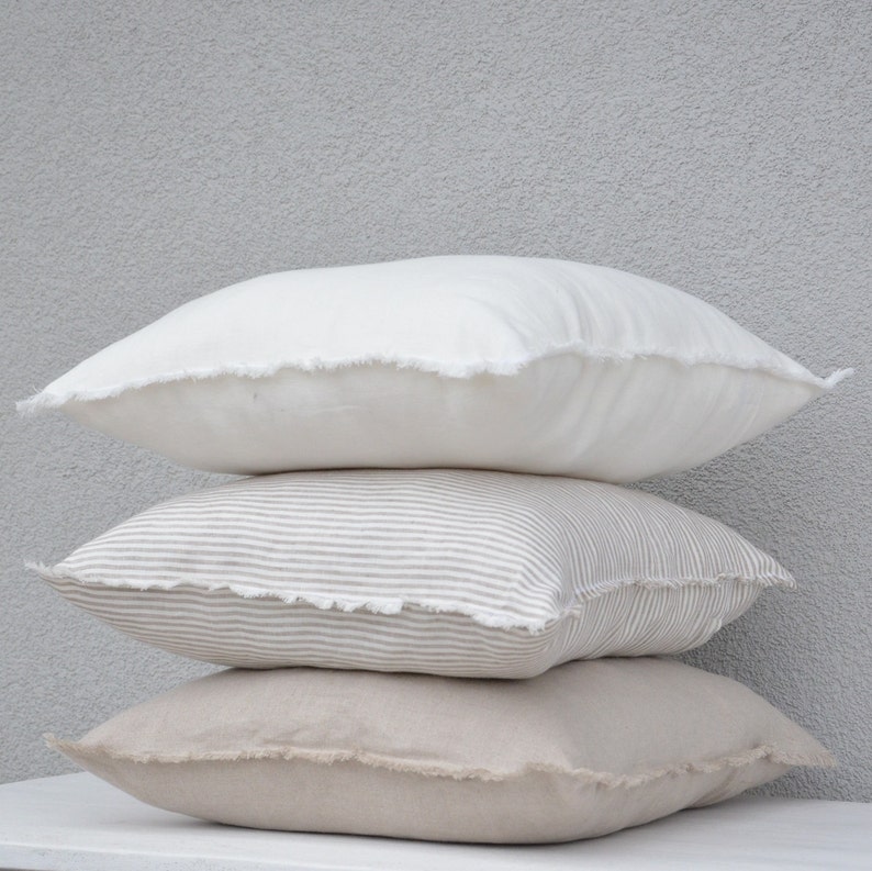 May include: Three decorative pillows stacked on top of each other. The top pillow is white, the middle pillow is striped in beige and white, and the bottom pillow is beige.