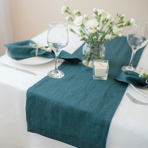 Puede incluir: Un camino de mesa de lino color teal con servilletas blancas y una vela blanca sobre un mantel blanco. La mesa está puesta con dos copas de vino y un ramo de flores blancas.