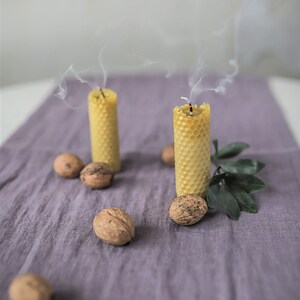 May include: Two honeycomb candles are burning on a purple linen tablecloth. The candles are made of beeswax and have a natural, rustic look. There are several walnuts scattered around the candles.