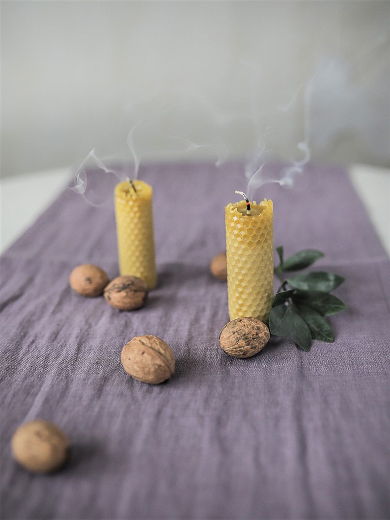 May include: Two honeycomb candles are burning on a purple linen tablecloth. The candles are made of beeswax and have a natural, rustic look. There are several walnuts scattered around the candles.