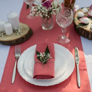 May include: A white linen tablecloth with a coral runner. A white plate with a coral napkin and small white flowers is set with a silver fork and knife. A glass of water and a small vase of white flowers are on the table.
