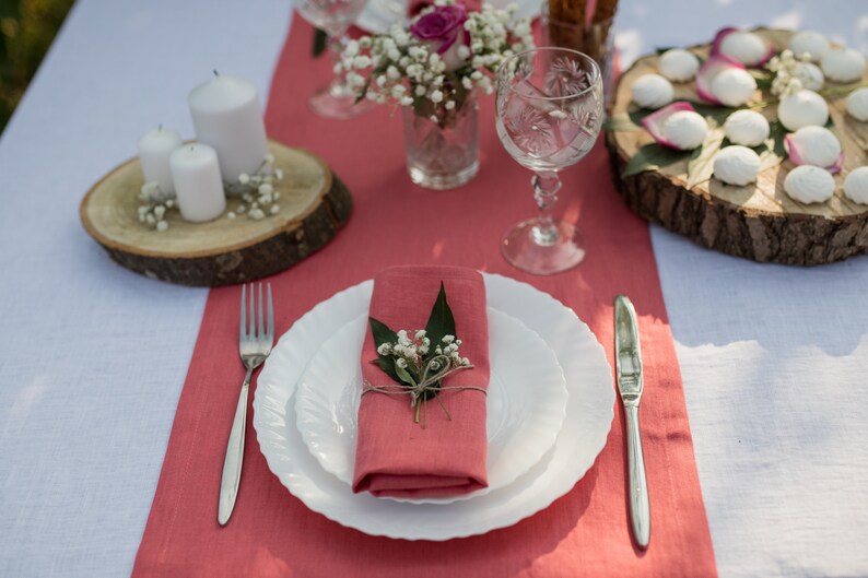 May include: A white linen tablecloth with a coral runner. A white plate with a coral napkin and small white flowers is set with a silver fork and knife. A glass of water and a small vase of white flowers are on the table.