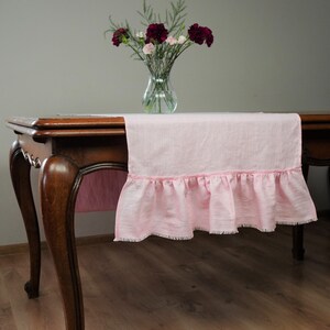 Pode incluir: Um corredor de mesa de linho rosa com uma borda franjada e uma borda desfiada. O corredor de mesa está sobre uma mesa de madeira com um vaso de flores rosa e bordô.