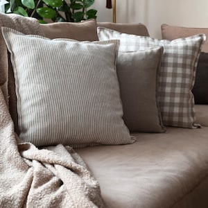 Pode incluir: Uma coleção de almofadas decorativas em tons neutros em um sofá bege. As almofadas incluem uma almofada listrada, uma almofada lisa e uma almofada com estampa xadrez. Uma manta bege está sobre o sofá.