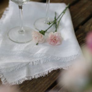 May include: Two white linen napkins with frayed edges, folded and placed on a wooden table. Two wine glasses are on top of the napkins, and two pink carnations are placed on the napkins.