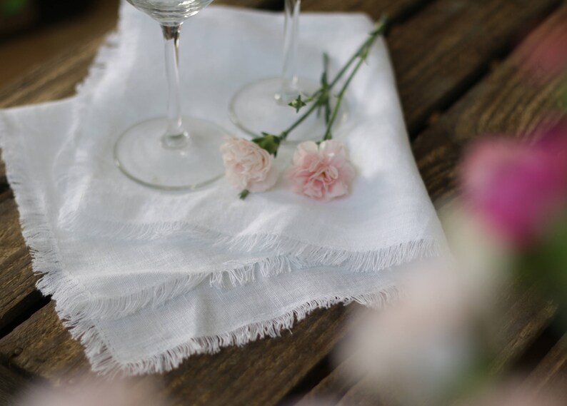 May include: Two white linen napkins with frayed edges, folded and placed on a wooden table. Two wine glasses are on top of the napkins, and two pink carnations are placed on the napkins.