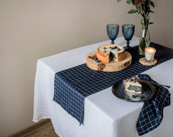 May include: A white tablecloth with a blue and white checked table runner and a blue and white checked napkin. A slice of poppy seed cake with blueberries on top is on a black plate. A whole poppy seed cake with blueberries on top is on a wooden chopping board. Two clear glass wine glasses and a candle are on the table.