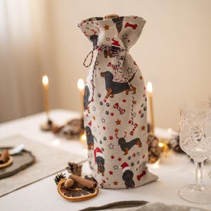 Könnte beinhalten: Eine beige Stoff-Weinflaschen-Tasche mit Kordelzug. Die Tasche ist mit einem sich wiederholenden Muster aus schwarzen und braunen Dackeln verziert, die Weihnachtsmützen tragen, rote und weiße Zuckerstangen und weiße Sterne.