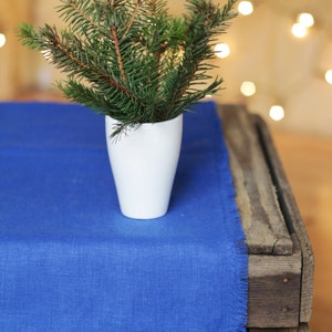 May include: A small sprig of pine needles in a white vase on a blue linen table runner. The table runner is on a wooden crate.