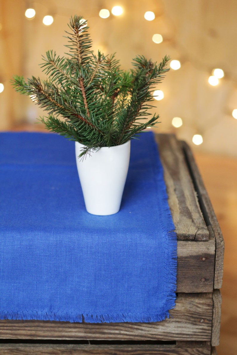 May include: A small sprig of pine needles in a white vase on a blue linen table runner. The table runner is on a wooden crate.