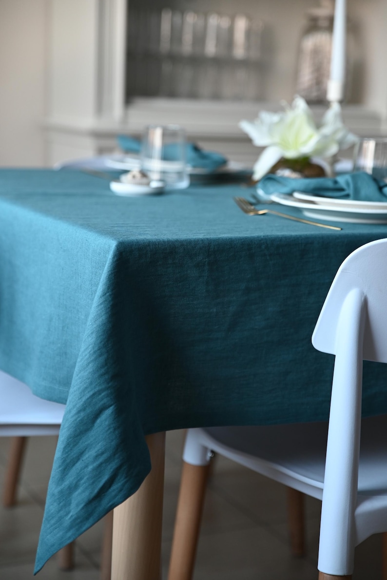 May include: Teal linen tablecloth draped over a wooden table, set for a meal. White chairs are positioned around the table. The table is set with white plates, teal napkins, and gold cutlery. A white flower arrangement is in the background.