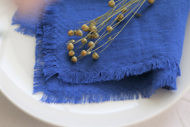 Pu&ograve; includere: Un tovagliolo di lino blu con un bordo sfrangiato, piegato e posizionato su un piatto bianco. Un rametto di fiori di lino secchi &egrave; posizionato sopra il tovagliolo.