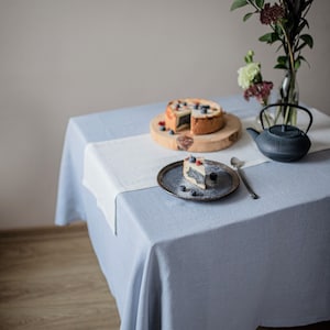 Op de afbeelding: Een lichtblauw linnen tafelkleed met een witte loper, gedekt voor een maaltijd. Een cake met bosbessen staat op een houten plank. Een stuk cake ligt op een klein bord. Een zwarte theepot en een vaas met bloemen maken de scène compleet.