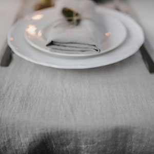 May include: A white dinner plate with a smaller white plate on top. A folded linen napkin is placed on top of the smaller plate, with a sprig of greenery tied around it. The plates are on a gray linen table runner.