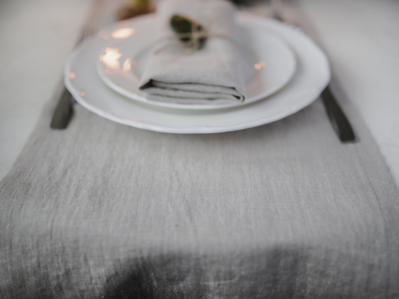 May include: A white dinner plate with a smaller white plate on top. A folded linen napkin is placed on top of the smaller plate, with a sprig of greenery tied around it. The plates are on a gray linen table runner.
