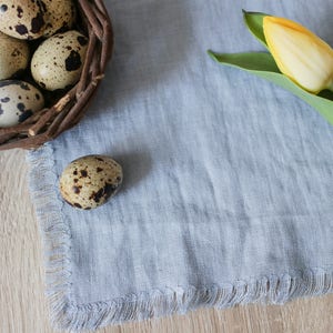 Pu&ograve; includere: Un cesto intrecciato pieno di uova di quaglia macchiate, con un solo uovo appoggiato su un tovagliolo di lino blu chiaro accanto a un tulipano giallo.