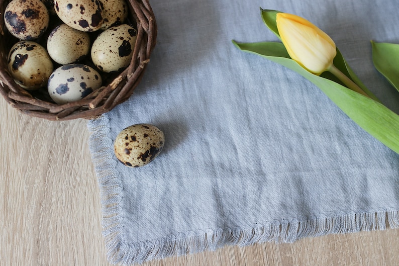 Pu&ograve; includere: Un cesto intrecciato pieno di uova di quaglia macchiate, con un solo uovo appoggiato su un tovagliolo di lino blu chiaro accanto a un tulipano giallo.