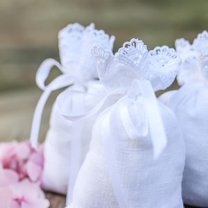 May include: Close-up of three white linen bags tied with white ribbons and adorned with delicate lace. Pink flowers are in the foreground. The bags are likely filled with potpourri or other scented items.
