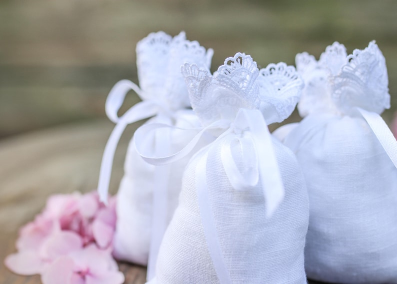 May include: Close-up of three white linen bags tied with white ribbons and adorned with delicate lace. Pink flowers are in the foreground. The bags are likely filled with potpourri or other scented items.