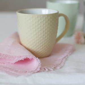 Pu&ograve; includere: Una tazza in ceramica di colore giallo pallido con una superficie testurizzata e un manico. La tazza &egrave; appoggiata su un tovagliolo rosa con frange su una tovaglia bianca.