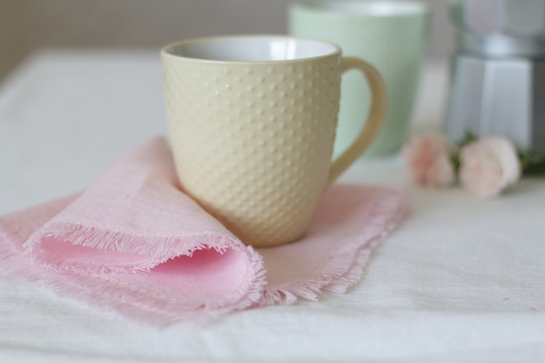 Pu&ograve; includere: Una tazza in ceramica di colore giallo pallido con una superficie testurizzata e un manico. La tazza &egrave; appoggiata su un tovagliolo rosa con frange su una tovaglia bianca.