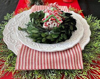 May include: A festive table setting with a red runner, white plates, and red and white striped napkins. A small gingerbread house sits atop a green wreath, surrounded by greenery and candles, creating a holiday atmosphere.