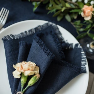 Pu&ograve; includere: Un tovagliolo di lino blu navy con bordi sfrangiati &egrave; piegato a forma di triangolo e posizionato su un piatto bianco. Un singolo garofano color pesca viene utilizzato come anello per tovagliolo.