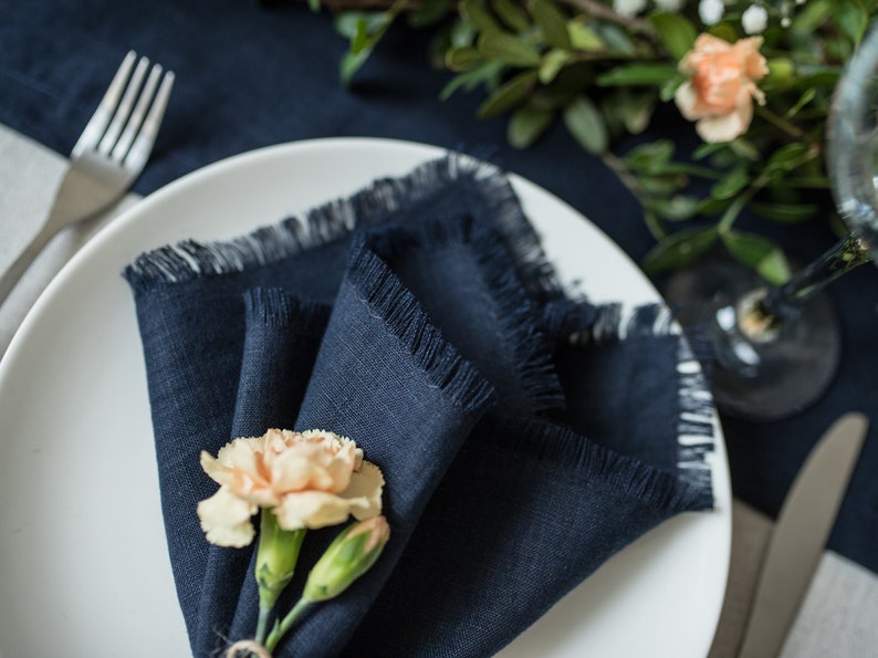 Pu&ograve; includere: Un tovagliolo di lino blu navy con bordi sfrangiati &egrave; piegato a forma di triangolo e posizionato su un piatto bianco. Un singolo garofano color pesca viene utilizzato come anello per tovagliolo.