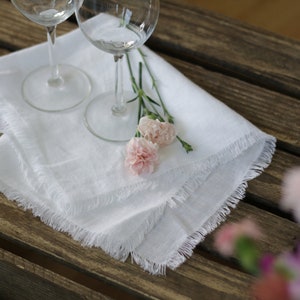 Pu&ograve; includere: Tovagliolo di lino bianco con bordi sfrangiati, due bicchieri da vino e garofani rosa su un tavolo di legno.