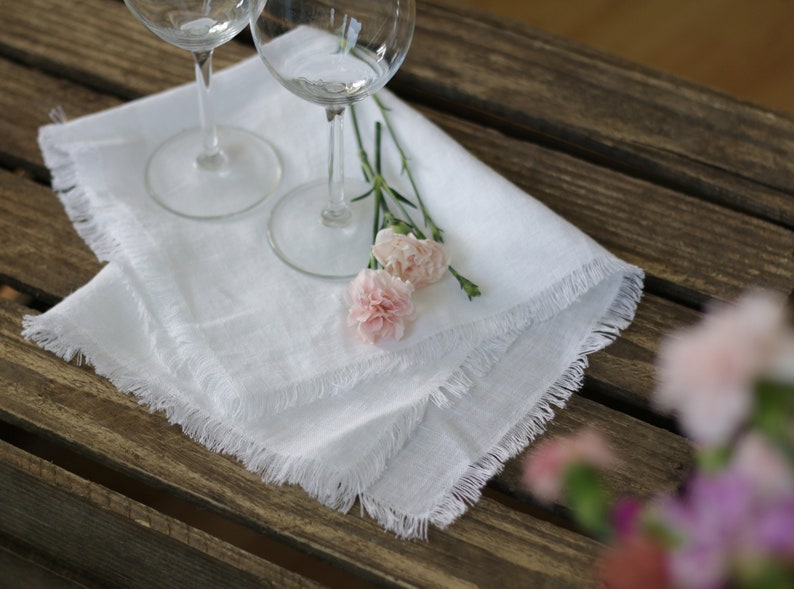 Pu&ograve; includere: Tovagliolo di lino bianco con bordi sfrangiati, due bicchieri da vino e garofani rosa su un tavolo di legno.