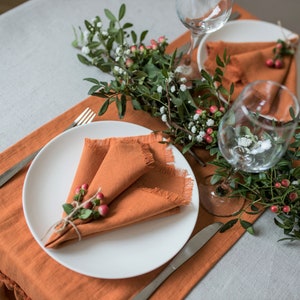 Pu&ograve; includere: Due coperti con piatti bianchi e tovaglioli di lino arancioni. I tovaglioli sono piegati a forma di triangolo e decorati con rametti di verde e bacche rosse. Un rametto di verde con bacche rosse corre lungo il centro del tavolo.