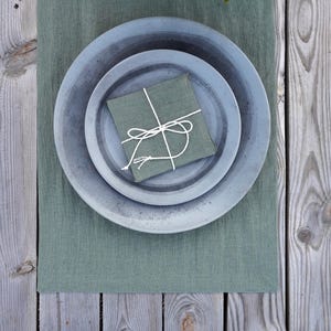 Op de afbeelding: Een set van drie grijze keramische borden met een rustieke afwerking, gestapeld op elkaar. Een groene linnen servet is vastgebonden met een wit touwtje en op de borden gelegd. De borden liggen op een groene linnen placemat.