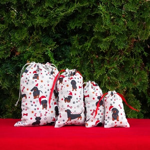May include: Four white fabric gift bags with a dachshund dog pattern. The bags are lined up in order of size, with the largest bag on the left and the smallest bag on the right. The bags have red ribbon ties and are sitting on a red surface.