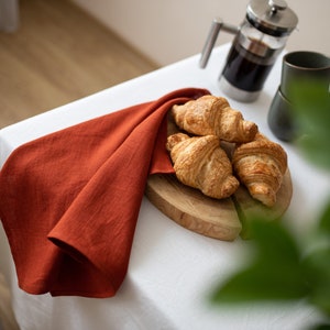 May include: Four golden brown croissants on a wooden cutting board with a red linen napkin on a white tablecloth.