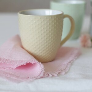 May include: A pale yellow ceramic mug with a textured surface sits on a pink fringed napkin. The mug has a handle and is partially visible against a white background.