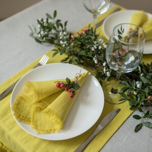 Pu&ograve; includere: Un piatto bianco con un tovagliolo di lino giallo piegato a forma di triangolo e legato con un rametto di verde e bacche rosse. Il piatto &egrave; posto su un sottopiatto di lino giallo con una forchetta e un coltello d'argento.