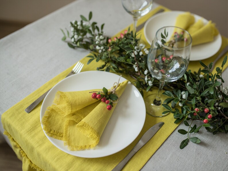Pu&ograve; includere: Un piatto bianco con un tovagliolo di lino giallo piegato a forma di triangolo e legato con un rametto di verde e bacche rosse. Il piatto &egrave; posto su un sottopiatto di lino giallo con una forchetta e un coltello d'argento.