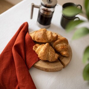 May include: Three golden brown croissants on a wooden cutting board with a red linen napkin in the background.