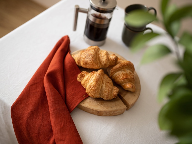 May include: Three golden brown croissants on a wooden cutting board with a red linen napkin in the background.