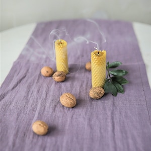 May include: Two yellow honeycomb candles are burning on a purple linen tablecloth. There are several walnuts scattered around the candles.